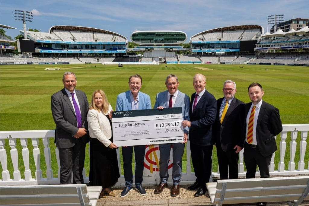 A group of seven people, including representatives from Tees and Synonymous Sport, hold a large charity cheque for £10,294.13 to Help for Heroes. They are standing in front of the cricket pitch at Lord’s Cricket Ground, with the iconic Pavilion and media centre visible in the background.