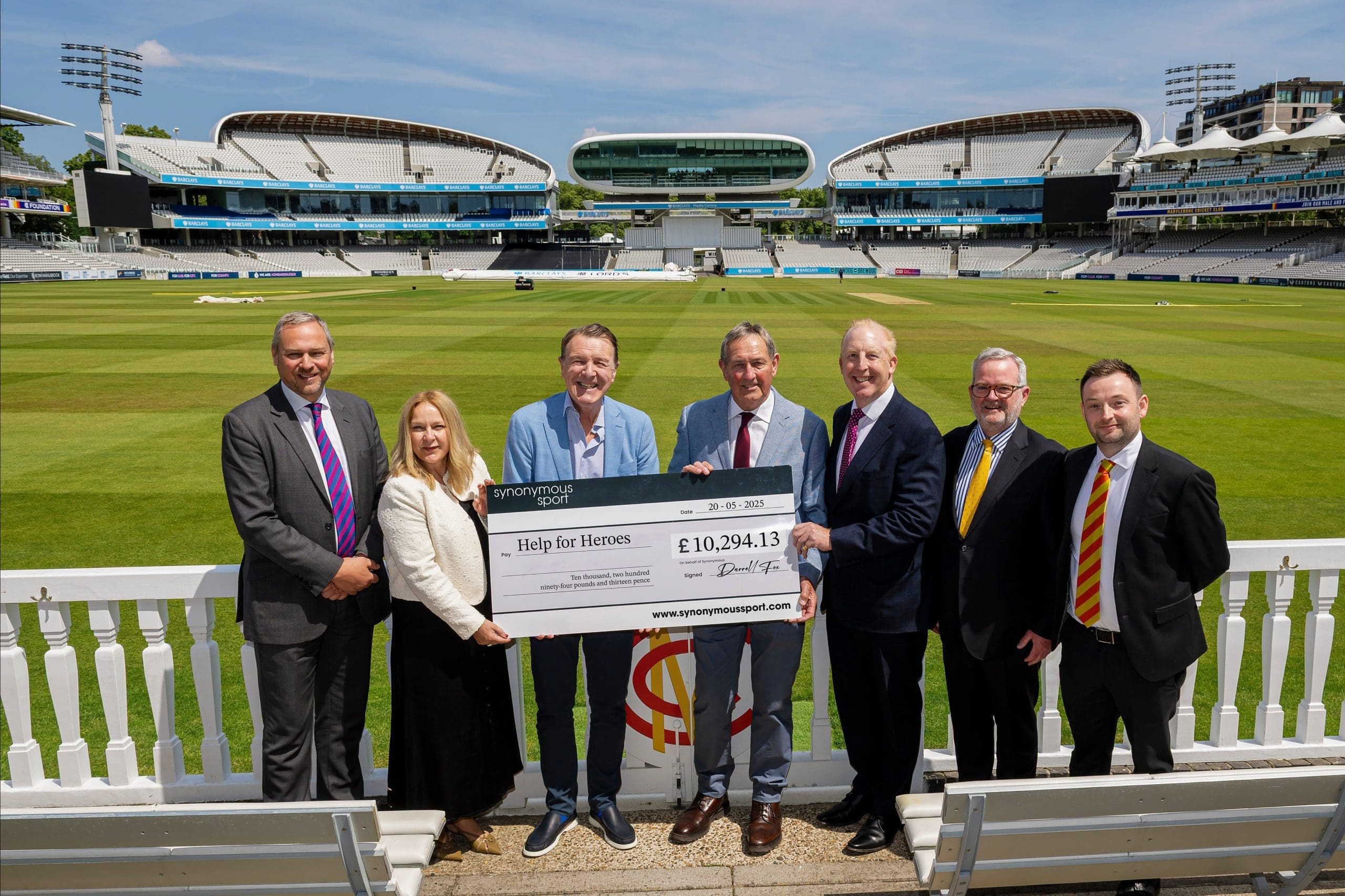 A group of seven people, including representatives from Tees and Synonymous Sport, hold a large charity cheque for £10,294.13 to Help for Heroes. They are standing in front of the cricket pitch at Lord’s Cricket Ground, with the iconic Pavilion and media centre visible in the background.