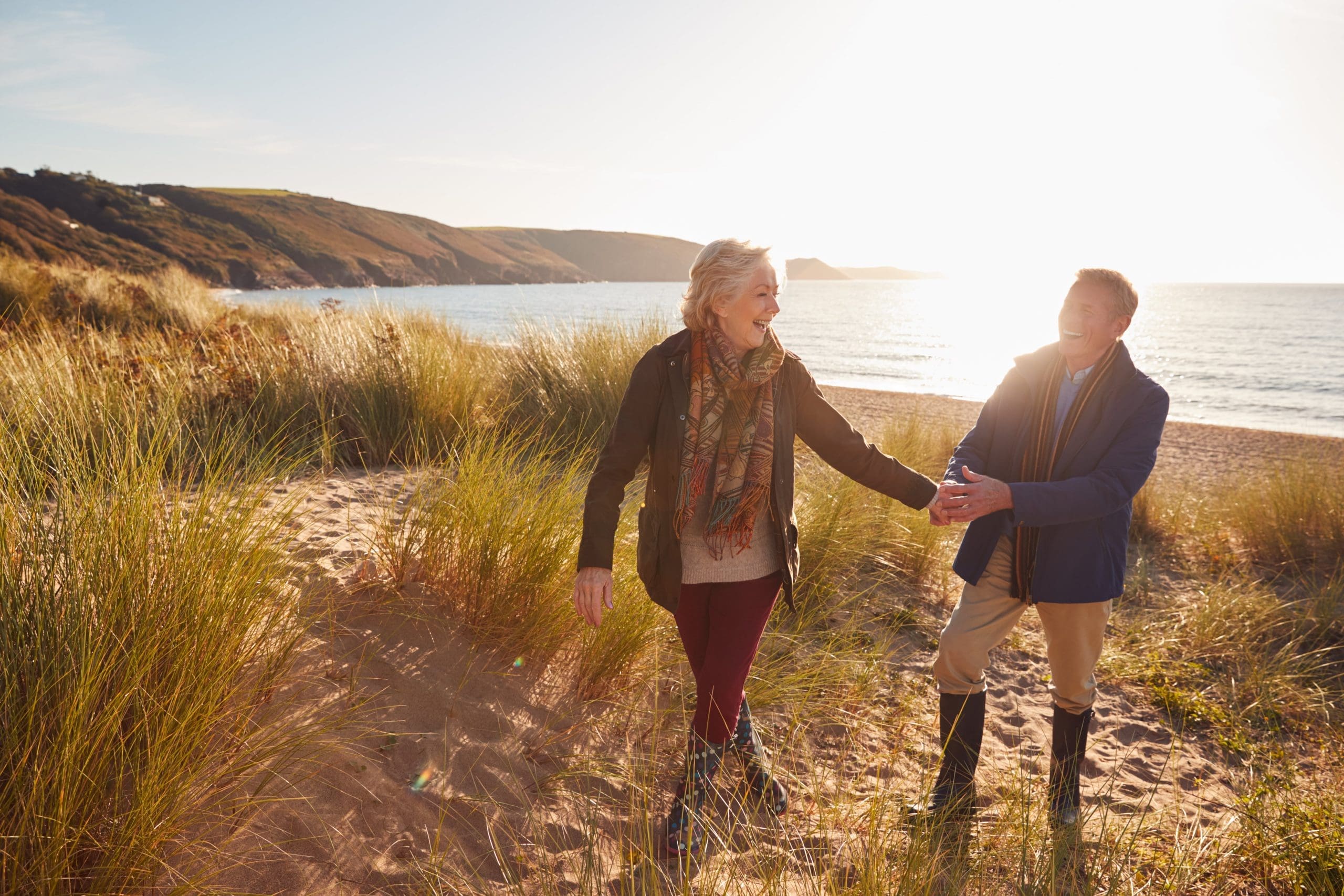 Older couple walking hand in hand on a beach at sunset, enjoying retirement.
