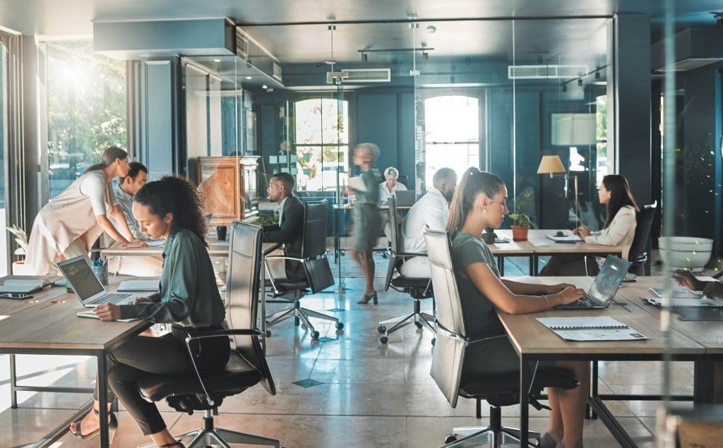 An open-plan office where several professionals are working at desks and in small groups, engaged in different tasks.