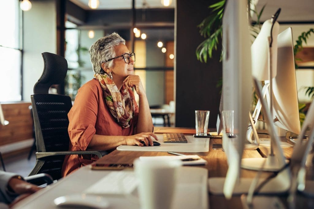 A businesswoman in an orange blouse and colorful scarf sits at a desk in a modern office, thoughtfully examining her computer screen. The workspace is neat and organized with multiple monitors and plants in the background, highlighting a focused and professional approach to business.