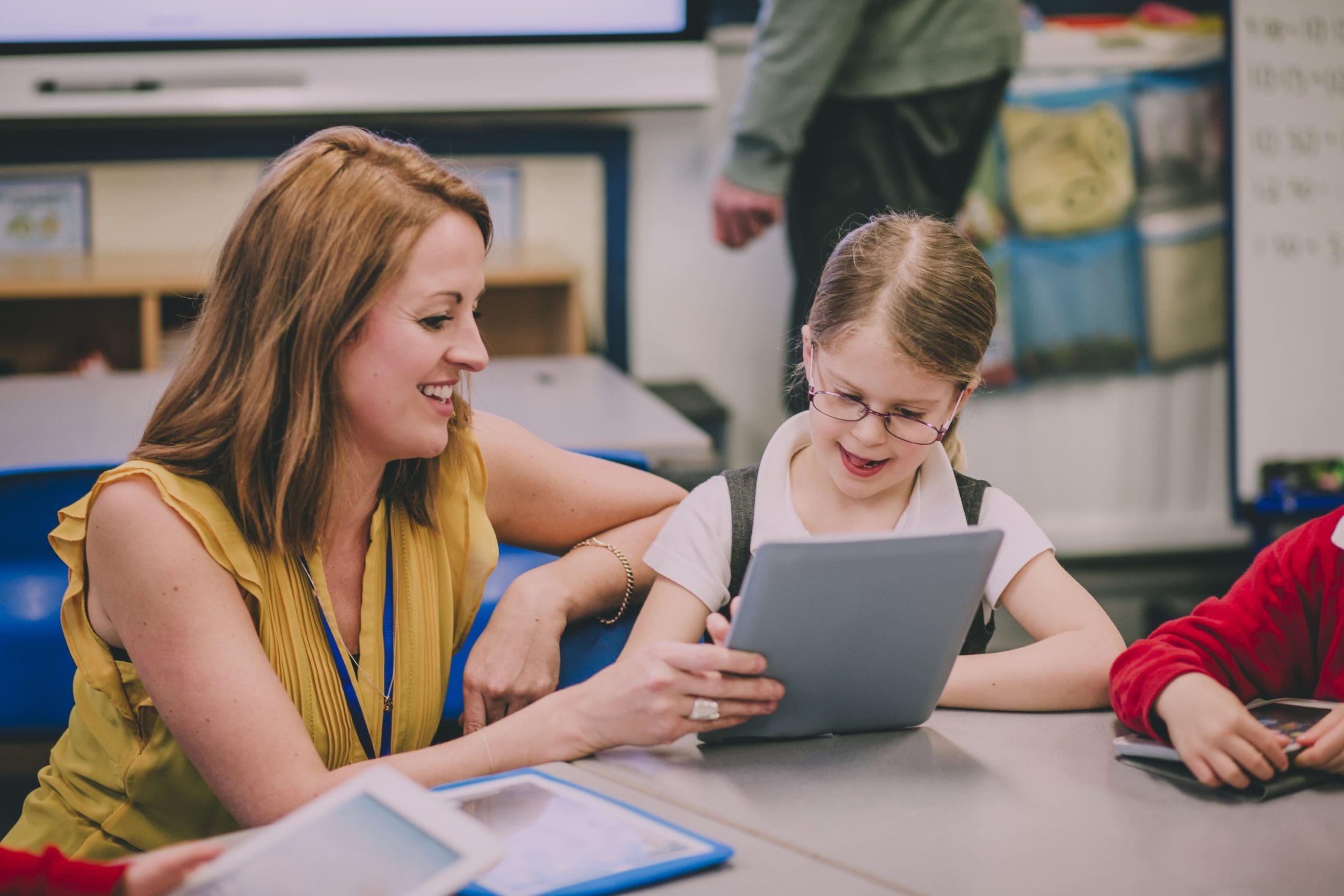 Teaching assistant supporting a young student with a tablet in a primary school classroom.
