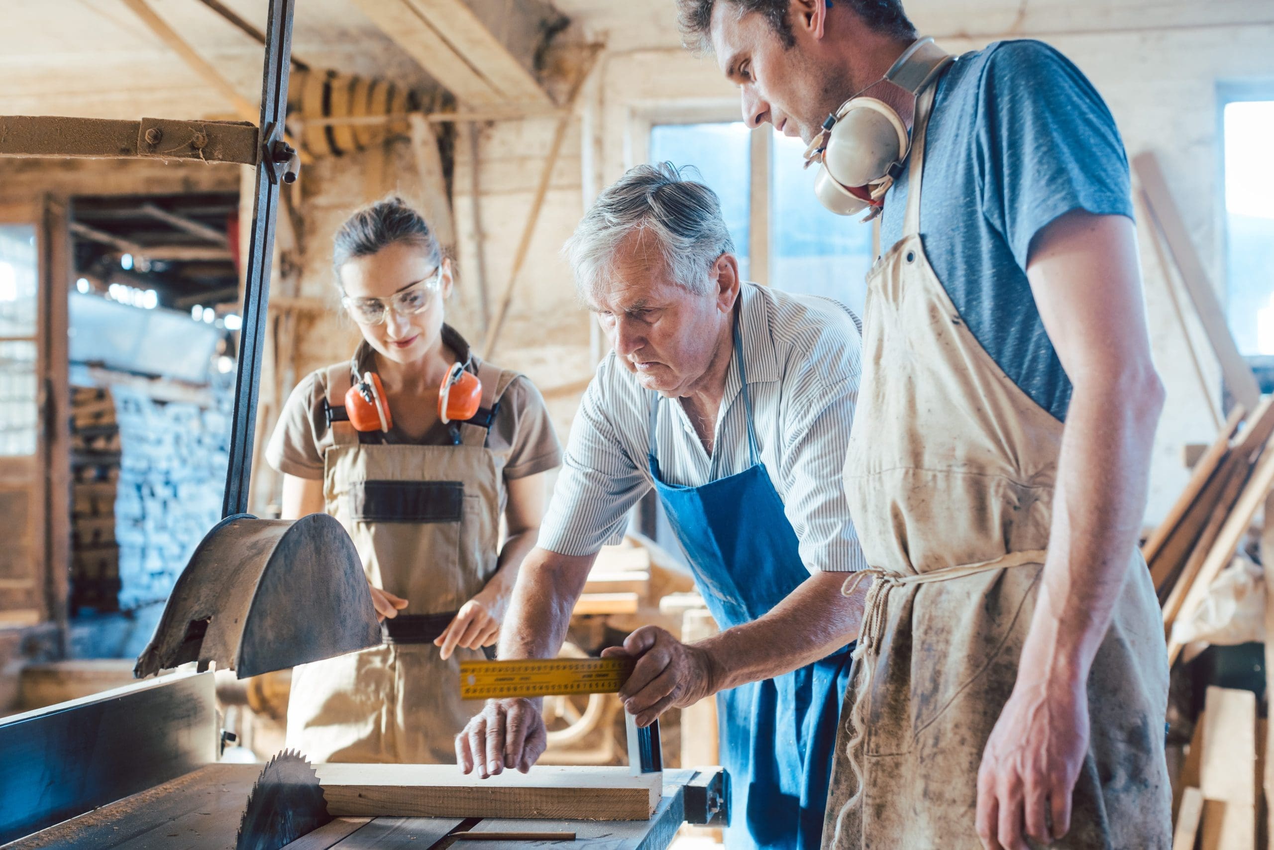 A family of three working together in a workshop, with the older man guiding the younger man and woman in woodworking tasks. They are all wearing protective gear, including safety glasses and earmuffs.