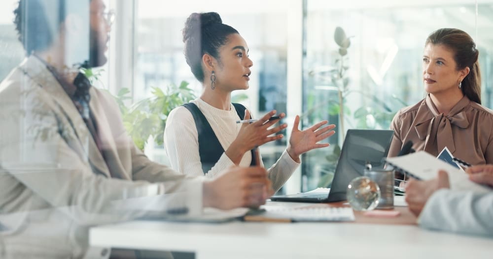 Four business professionals in a modern office having a serious discussion, with one woman gesturing while explaining a point to her colleagues.