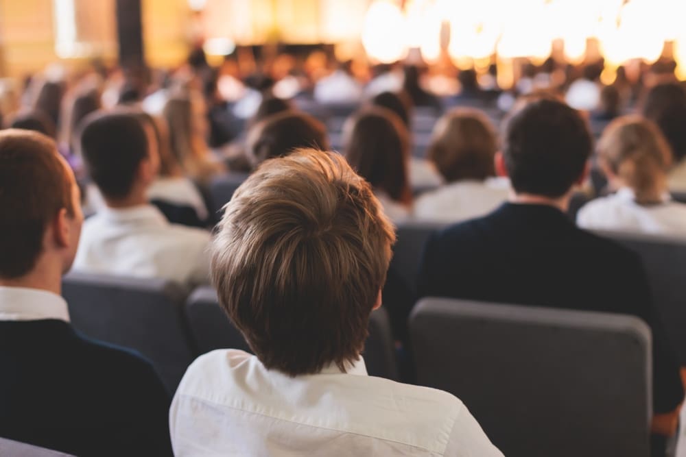 Students seated in a large school assembly hall facing a stage, representing school events where crowd safety and emergency planning are important under Martyn’s Law.