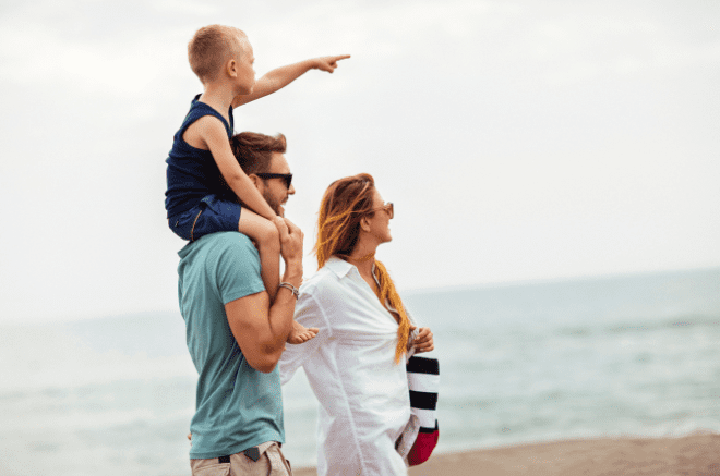 a woman man and child walk along a beach, the child is on the mans shoulders and is pointing ahead